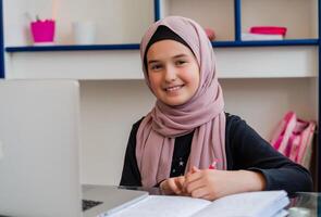 Portrait of a happy female Muslim student in hijab smiling and looking at the camera, sitting at a desk using a laptop computer and learning for school in the classroom or at home. photo