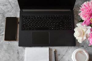 On the left side of the table lies a black smartphone, and a white disposable cup with a lidOn the right side of the table, a bouquet of pink and white peonies is visible. photo