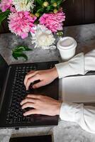 A woman is sitting at a table and typing on a laptop.A bouquet of peonies is placed on the table behind the laptop. photo