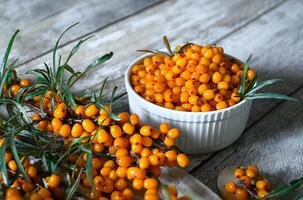 A photo of a white wooden table with an arrangement of ingredients for a healthy and flavorful drink, including sea buckthorn berries, lemon slices, honey, and cloves.