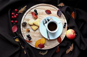 There is a cup of coffee, a saucer, a spoon, three cookies, an apple, and various autumn leaves on the tray. photo