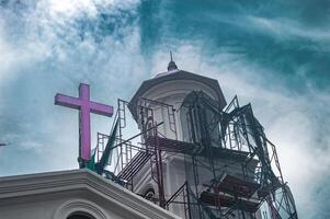 A church undergoing renovation with scaffolding surrounding its tower and a prominent purple cross against a cloudy sky, Indonesia, 16 November 2024 photo