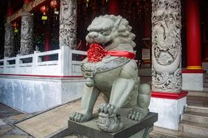 A stone lion statue adorned with a red ribbon stands guard at the entrance of a temple, featuring intricate carvings and festive decorations for the Lunar New Year, Indonesia, 17 November 2024 photo