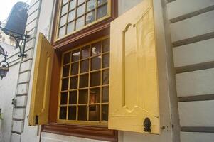 A vintage window with yellow shutters open, showcasing a grid patterned glass pane. The scene includes a decorative wall lamp and a white exterior wall, evoking a historical or colonial ambiance photo