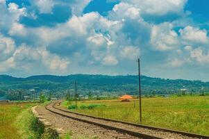 Scenic railway track curving through lush green fields with a backdrop of rolling hills and a vibrant blue sky filled with fluffy clouds. Perfect for travel and nature themes photo