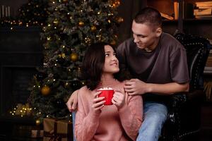 Embracing the New Year spirit, a couple in love sips hot cocoa with marshmallows, nestled beside a charming tree adorned with gold toys photo