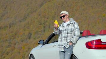 Successful happy guy watches at smartphone stands near whate car in mountains. video