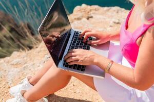 A woman is sitting on a rock with a laptop open in front of her. She is typing on the keyboard and she is focused on her work. Concept of productivity and concentration. photo