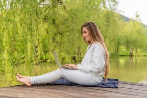 A woman is sitting on a dock with a laptop open in front of her. She is focused on her work, possibly typing or browsing the internet. Concept of productivity and concentration. photo
