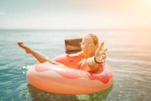 Woman laptop sea. Freelancer woman in sunglases floating on an inflatable big pink donut with a laptop in the sea. People summer vacation rest lifestyle concept photo