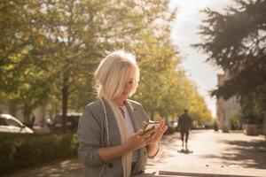 A blonde woman is looking at her cell phone while walking down a sidewalk. The scene is sunny and peaceful, with trees in the background. photo