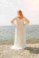 woman white dress stands on a beach, looking out at the ocean. She is in a contemplative or reflective mood, as she raises her hands above her head. Concept of peace and tranquility. photo