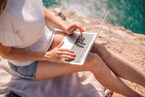 Freelance woman working on a laptop by the sea, typing away on the keyboard while enjoying the beautiful view, highlighting the idea of remote work. photo