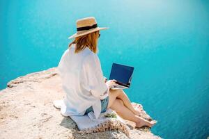 Freelance woman working on a laptop by the sea, typing away on the keyboard while enjoying the beautiful view, highlighting the idea of remote work. photo