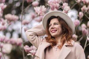 Woman magnolia flowers, surrounded by blossoming trees., hair down, white hat, wearing a light coat. Captured during spring, showcasing natural beauty and seasonal change. photo