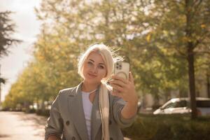 A woman is taking a picture of herself with her cell phone. She is wearing a gray jacket and scarf. The scene is set in a city with trees and cars in the background. photo