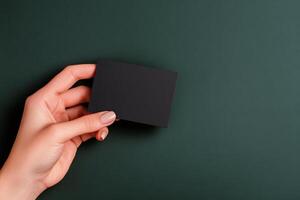 Hand holding a blank black card against a dark green background in a minimalist composition photo