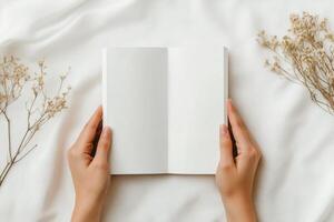 Hands holding an open blank book on a soft white surface with delicate flowers photo