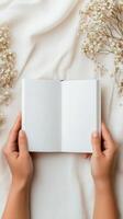 Open blank notebook held by hands on soft fabric with dried flowers nearby in natural light photo