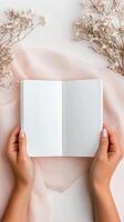 Open blank notebook held by hands on soft fabric with dried flowers nearby in natural light photo