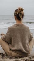 A woman with her hair tied back sits cross legged on the sandy beach, wearing a cozy sweater photo