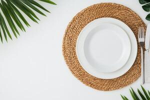 Clean white plate on a woven placemat surrounded by green foliage and cutlery photo