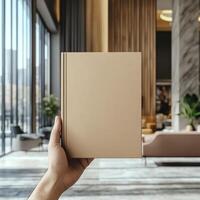 Hand holding a plain brown book in a stylish interior with greenery in the background photo