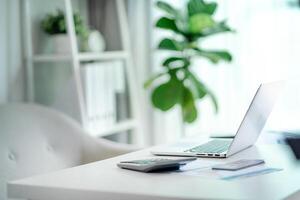 A white desk with a laptop, calculator, and a potted plant. The desk is clean and organized, with a sense of productivity and focus. The potted plant adds a touch of nature and freshness to the space photo