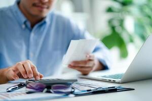 A man is sitting at a desk with a calculator and a receipt in front of him. He is focused on the receipt, possibly checking his finances or making a payment photo