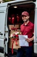 Smiling delivery man wearing red cap and T-shirt with documents and bouquet of roses in hands near the open side door of a white van with gift boxes, and heart-shaped balloons photo