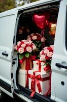 Open side door of a white delivery minivan with gift boxes with red ribbons, bouquets of roses, and a heart-shaped red balloons, close-up view photo