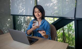 Asian woman sitting on chair holding mobile phone credit card in hand and looking at laptop for shopping online, internet banking and payment success, relaxing lifestyles concepts photo