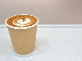side view beauty white foam design couple flower heart in brown paper cup of coffee on white table and brown wall background. save earth for use recycle container. photo