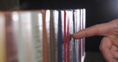 A mans hand is looking for the right book among a collection of volumes of books on a wooden bookshelf. Close Up. Shallow depth of field. Ultra 4K video