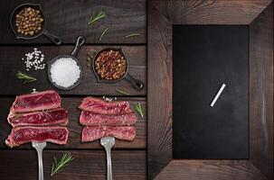 Juicy pieces of roast beef are skewered on a fork, spices. Nearby is an empty black chalk board for writing a menu photo