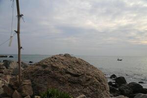 A large rock by the sea, with an iron pole on top of it and white paper hanging from both sides photo