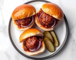 A top-down view of three meatballs glazed in red sauce, on buns with pickles and sliced onion, placed on a white plate against a clean background photo