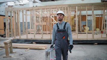 Worker carpenter in uniform walks with tools near unfinished prefabricated modular house in construction site video
