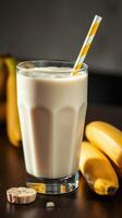 Creamy banana smoothie in a tall glass with yellow striped straw beside fresh bananas on a wooden table photo