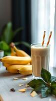Creamy banana smoothie served in a glass with yellow bananas in the background and tropical leaves around photo