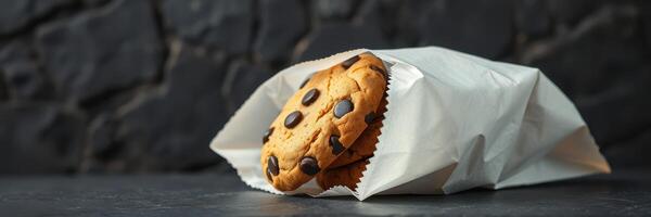 Warm chocolate chip cookie wrapped in paper on dark stone surface photo