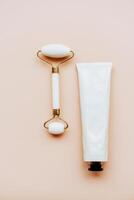 White jade facial roller and metallic cream tube with a black cap on a soft beige background photo