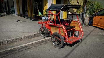 pedicab or rickshaw or locals calls it becak with a canopy is parked on a sunny street, reflecting a vibrant urban scene. The vehicle features a padded seat and unique design photo