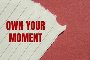 Own your moment message written on ripped torn lined paper with red background. Conceptual own your moment symbol. Copy space. photo