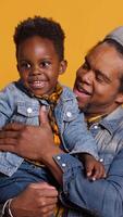 Vertical African american father and cute toddler having fun together in studio, dad carrying and embracing him. Young dad does good parenting with his infant, showing love and affection. Camera A. video