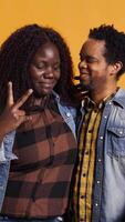 Vertical Cute life partners posing in studio and doing peace sign in studio, showing the victory symbol against yellow background. African american spouses doing kawaii gesture. Camera B. video