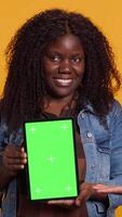 Vertical African american couple pointing at a tablet with isolated green screen display, smiling at each other against yellow background. Cheerful affectionate husband and wife posing. Camera A. video
