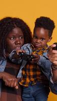 Vertical Cheerful african american dad playing games with his son on console, using controller to have fun against yellow background. Happy positive young family enjoying their leisure time. Camera A. video