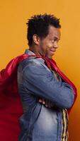 Vertical African american dad being the superhero of his little toddler, posing as a hero with a red cape and saving his family from any danger. Cute loving family posing against yellow background. Camera B. video