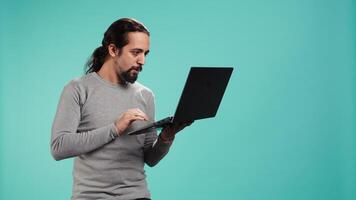 Upset man frowns while looking at laptop, dissatisfied with company profits, studio backdrop. Irked worker flabbergasted by low business income revenue, doing financial analysis on notebook, camera B video